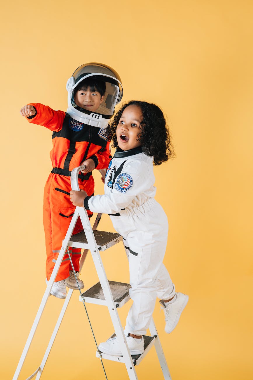 Children in astronaut uniforms on a step ladder pointing at something out of shot.