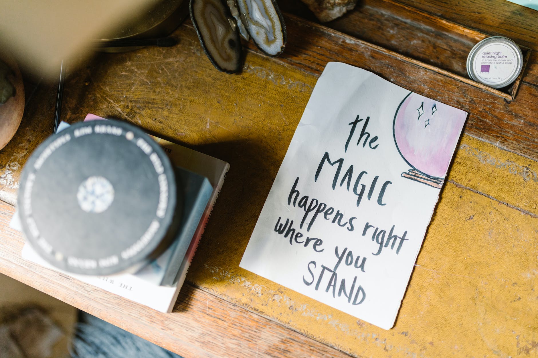 An old wooden desk with some clutter on it. On the top of the desk, seen from above, is a sign reading 'the MAGIC happens where you STAND'