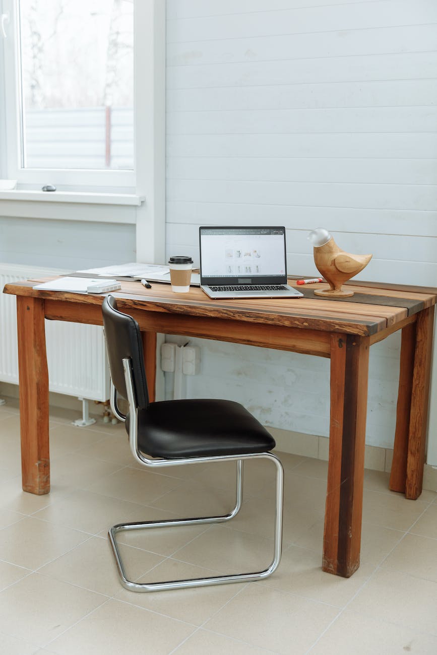 Desk with a laptop, coffee cip and a few items on it, next to a white painted wall with a window. Next to the desk is an empty chair.