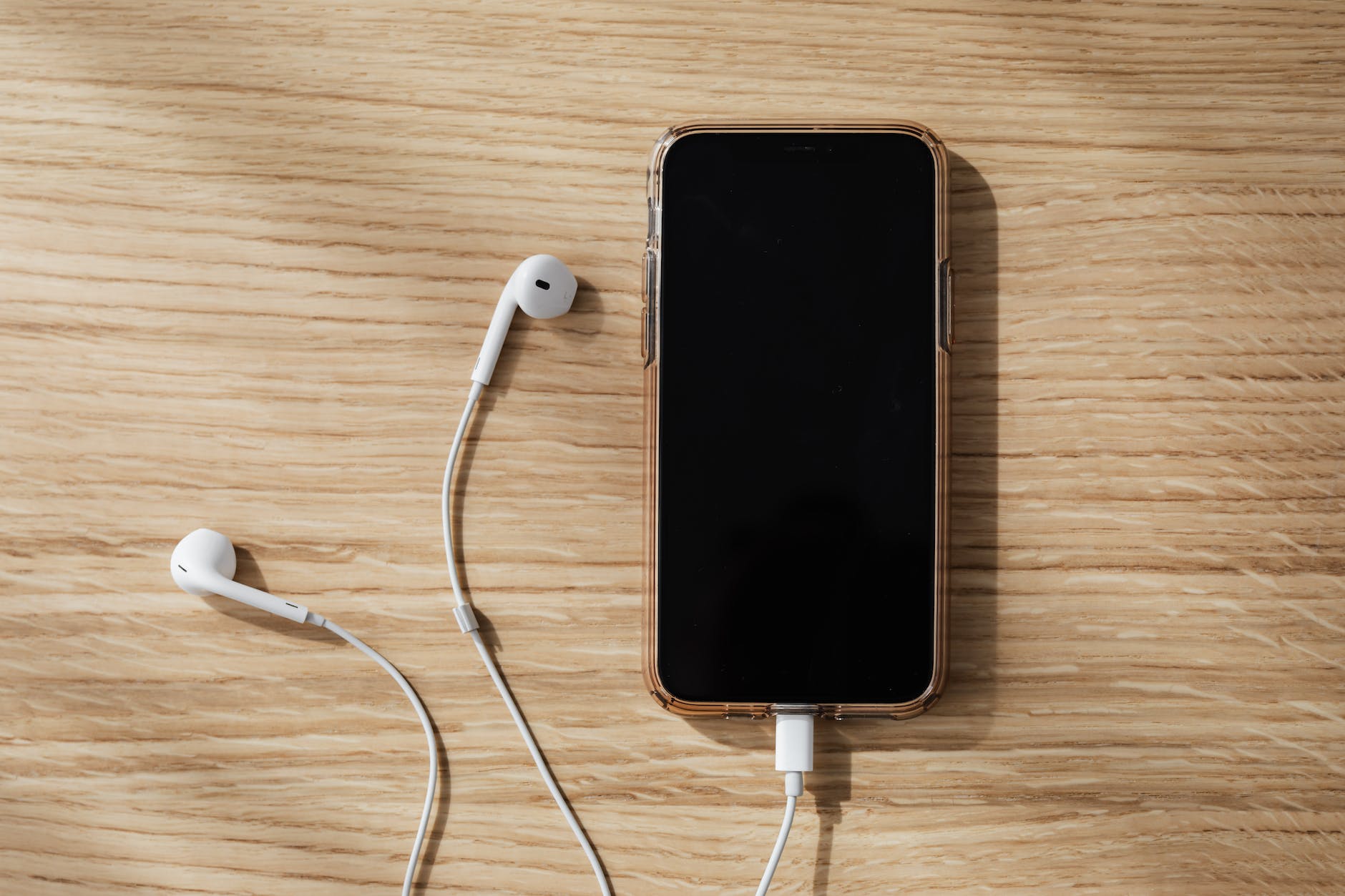A mobile phone on a wooden desk with white wired ear buds connected.