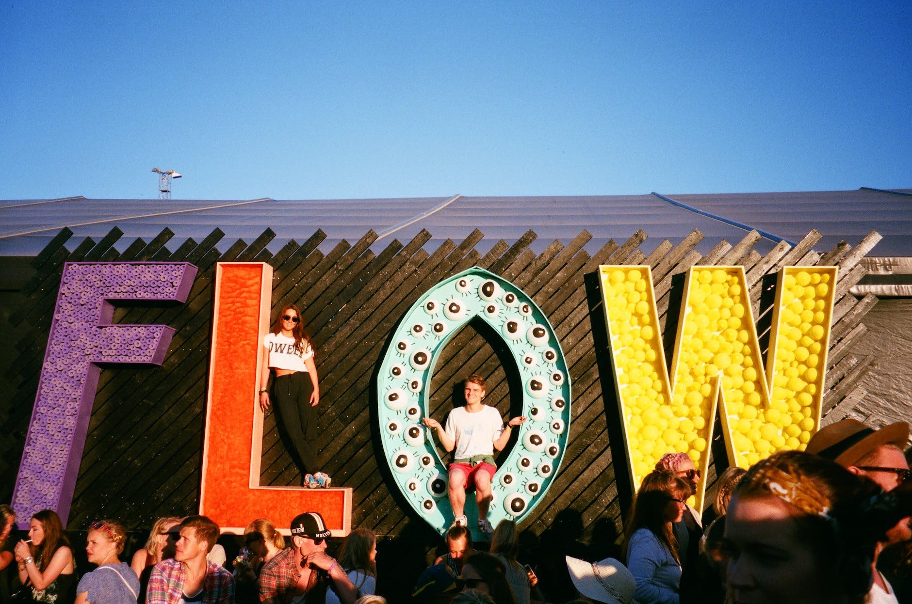 Large sign which spells 'FLOW'. A woman is standing on the 'L' and a man is sitting in the 'O'.