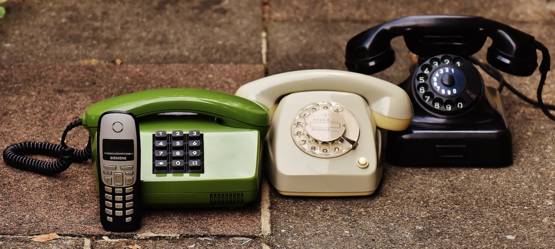 Four telephones - a vintage dial phone, a more modern dial phone, a push button landline phone, and an early press button mobile.