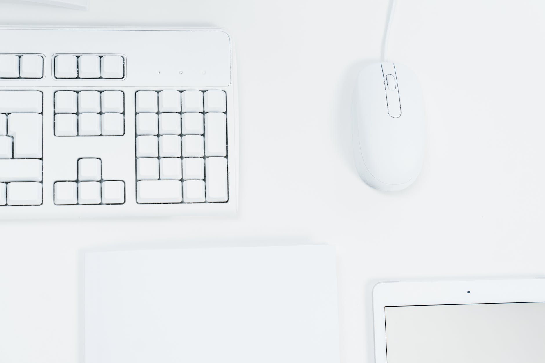 A white wired mouse, a white iPad, and a white keyboard pictured against a white desk.