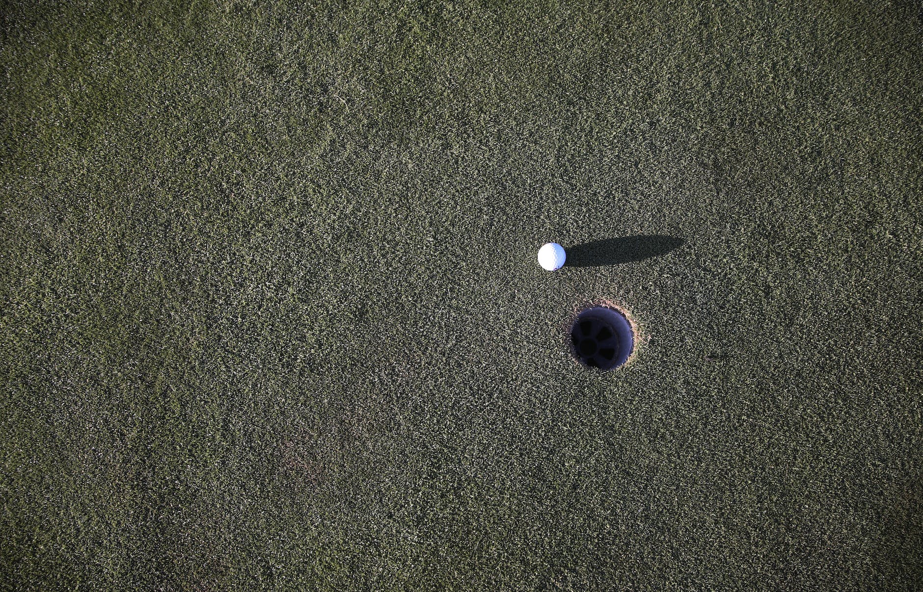 Picture of a golf ball near a golf hole, taken from above. The ball is casting a long shadow on the washed out grass.