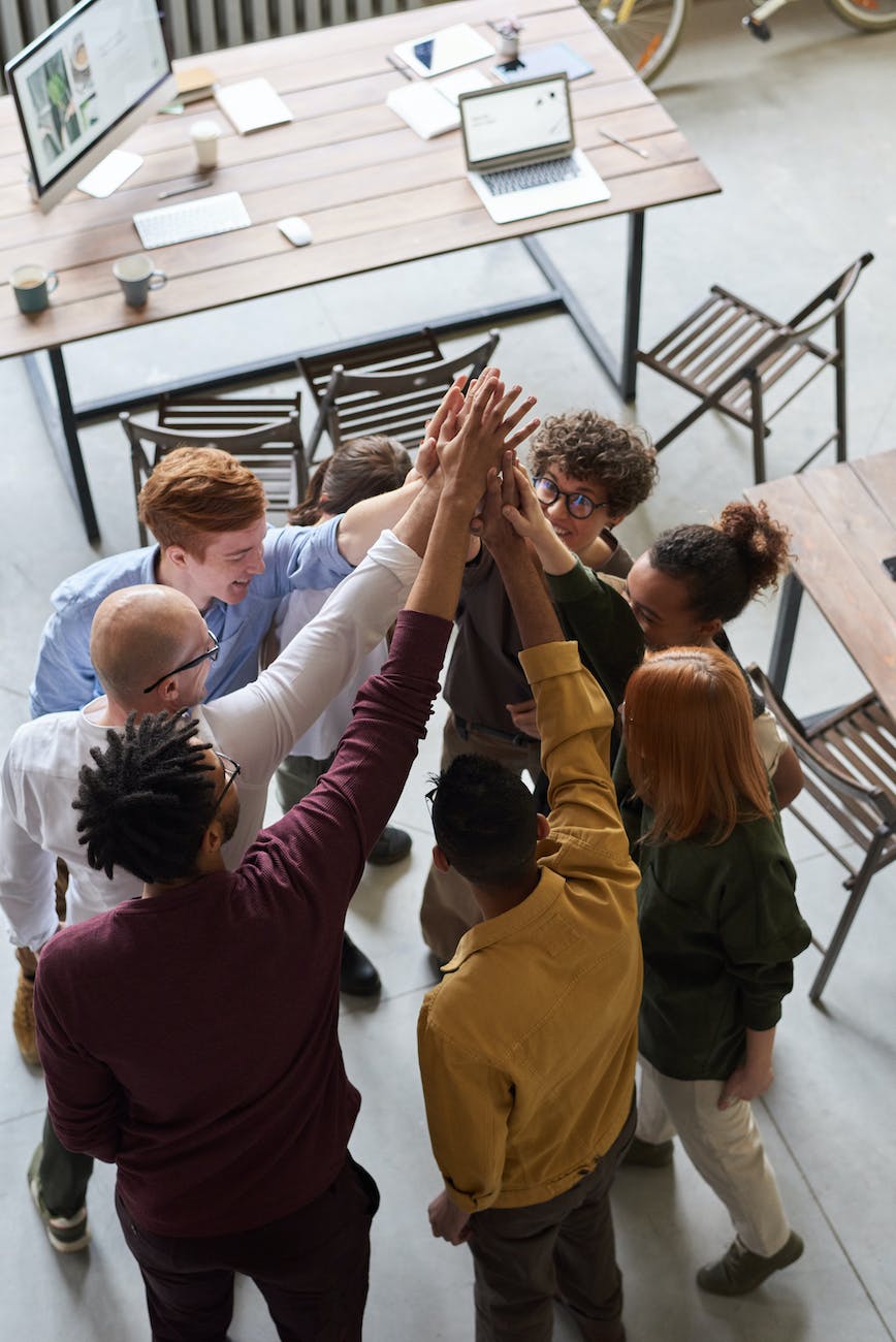 Clichéd picture of a diverse group of people standing in a circle in an office holding their hands up to the ceiling.
