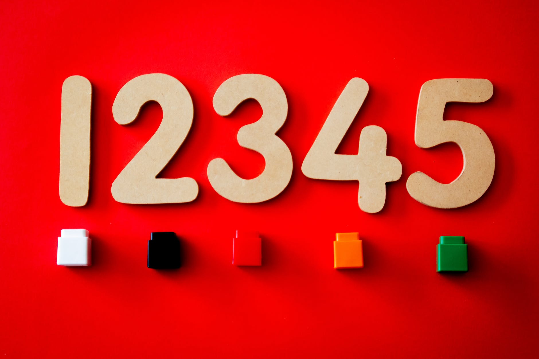 
The image depicts large wooden numbers from 1 to 5 arranged in order on a vibrant red background. Below the numbers, there are four small, square blocks in white, black, red, and orange, and a green rectangular block to the right. The contrast between the neutral colours of the numbers and the bright hues of the blocks creates a visually striking composition.