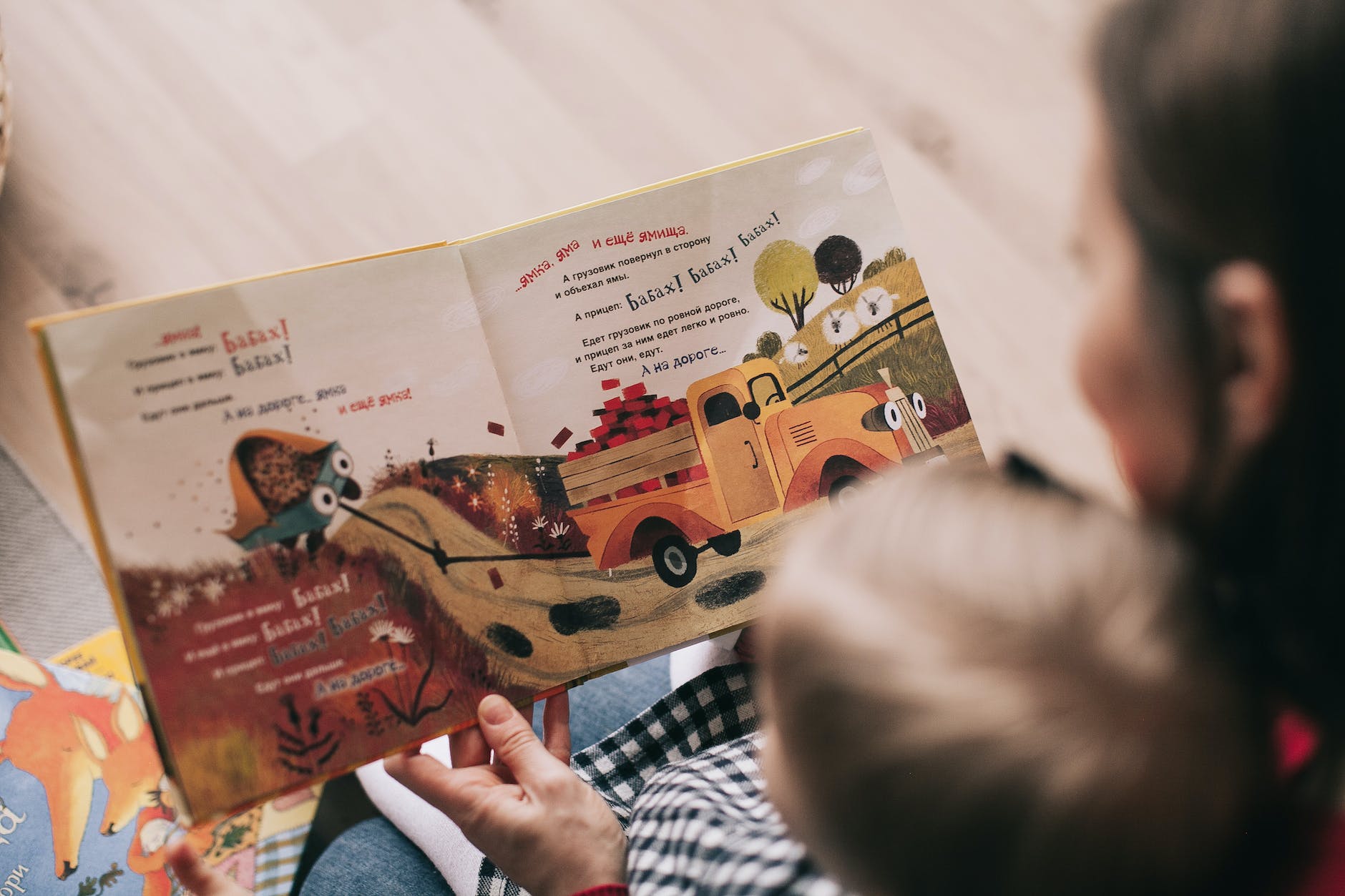 An adult reads a non-English illustrated book to a child, featuring a fire truck and animals, in a cozy indoor setting.