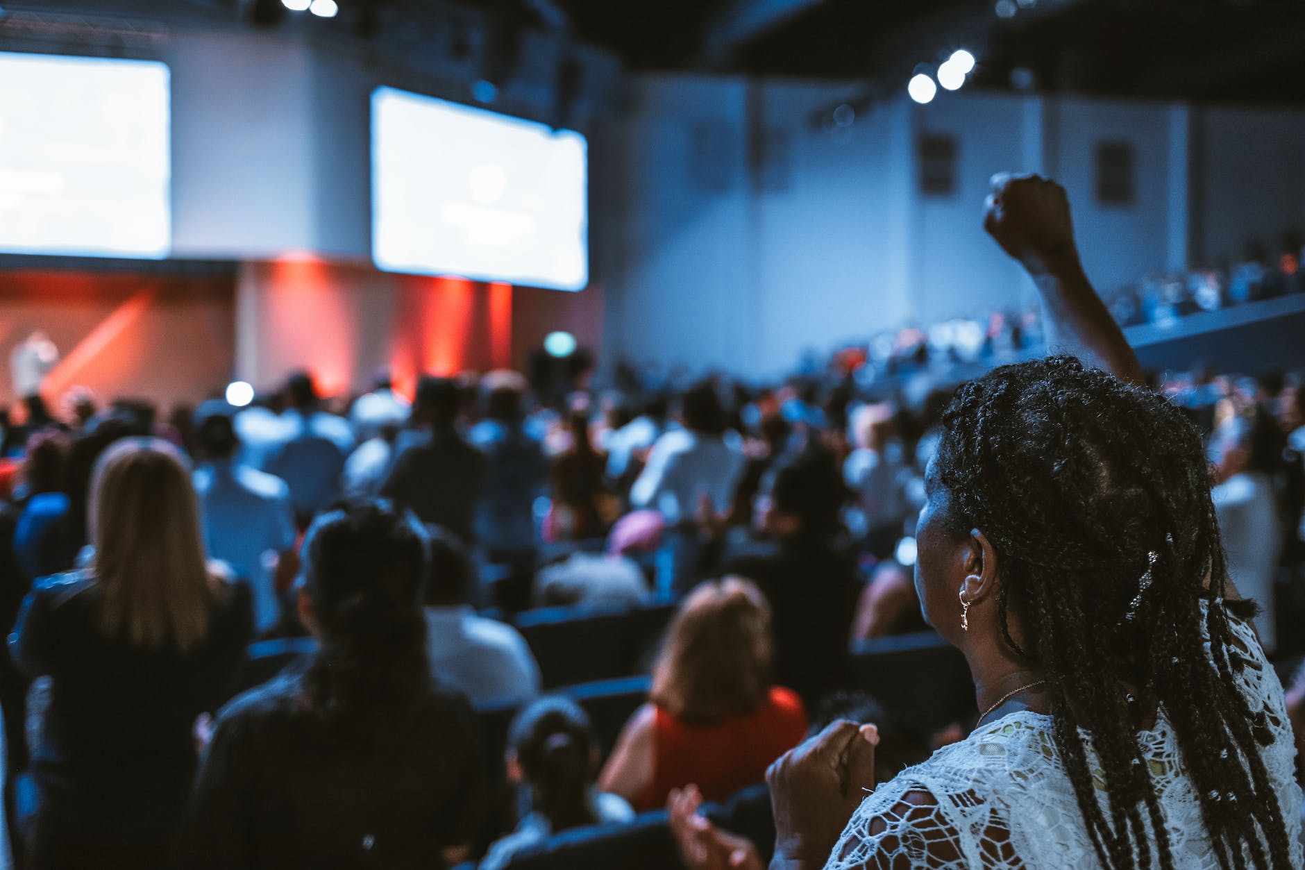 A woman raising her hand in a crowded auditorium, possibly participating or reacting to an event or speaker. She is in focus, with the audience in the background slightly blurred.