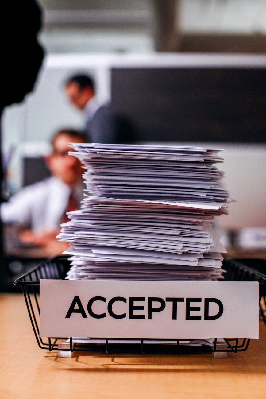 The image shows a large stack of documents neatly placed in a wire basket. On the front of the basket, there is a prominent label with the word "ACCEPTED" in bold, capitalized letters. The focus is on the stack and the label, with a blurred background featuring people in an office setting.