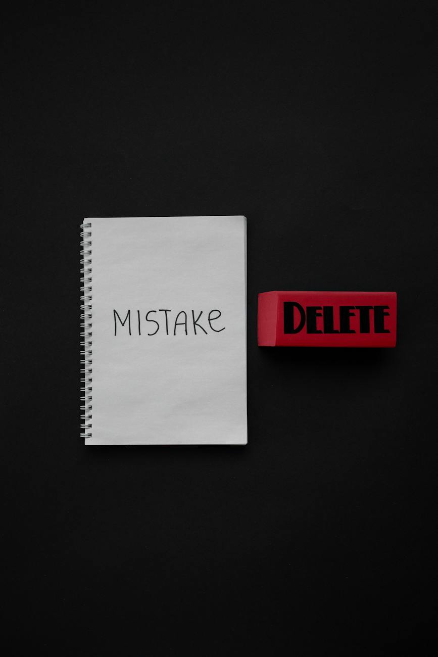 The image shows a notebook on a black background with the word "MISTAKE" written on it, alongside a red eraser with the word "DELETE" printed on it.