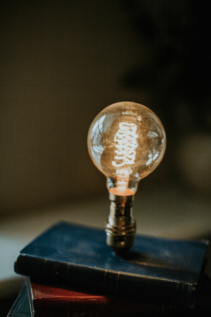 An illuminated light bulb stands on top of a stack of hardcover books in a dimly lit room, suggesting inspiration or ideas.