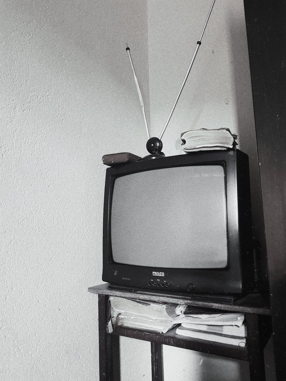 An old-fashioned television with rabbit ear antennas on top of a stack of newspapers on a wooden stand, in black and white.






