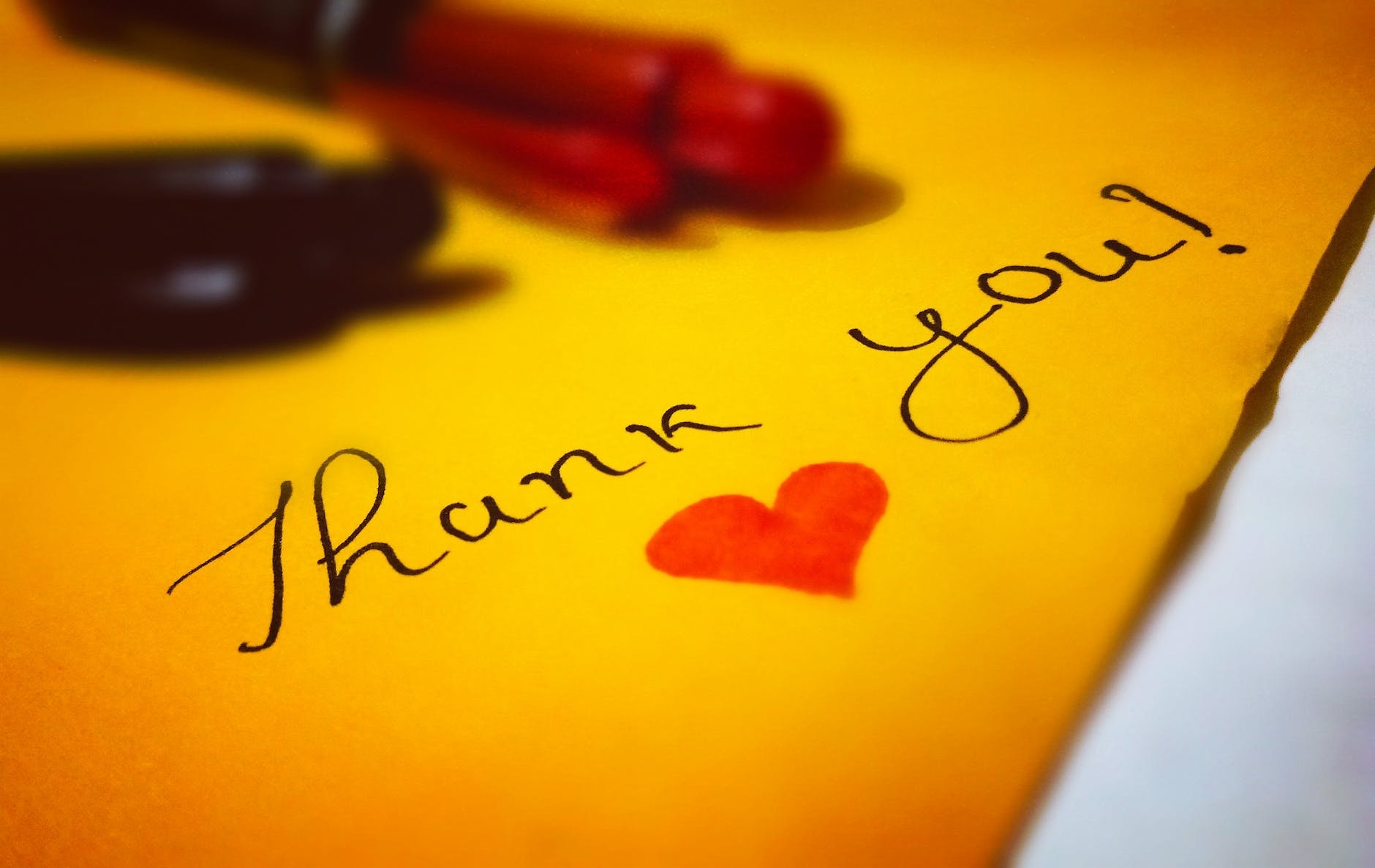 The image features a handwritten note saying "Thank you!" with a red heart symbol next to the text. The note is on a yellow surface, and there are blurry images of a pen and a part of another object in the foreground.
