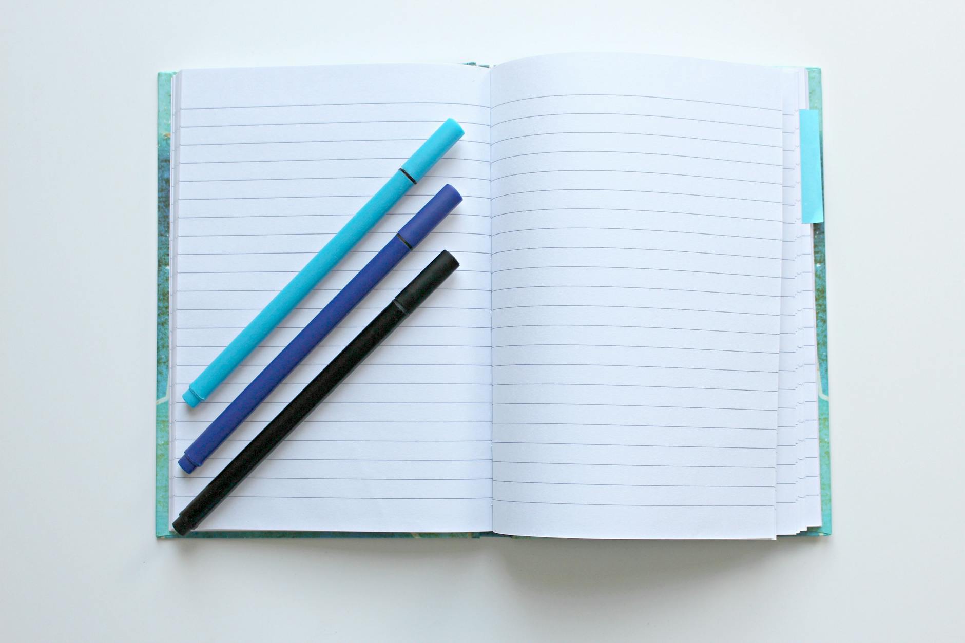 The image features an open lined notebook lying on a white surface. Three pens, one light blue, one dark blue, and one black, rest diagonally across the left page. The notebook's pages are white with blue horizontal lines. The notebook's cover has a marbled green and blue design, and the right page has a pale blue sticky note protruding from the top, serving as a makeshift bookmark.