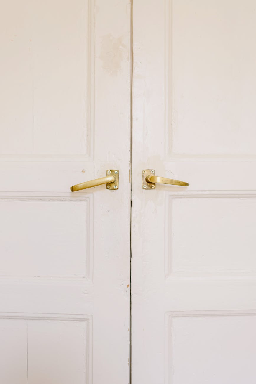 
This image displays a pair of white double doors that are closed. Each door has a panel design and features a brass-colored door handle mounted on the right side. The doors and handles show signs of wear and age, with some visible chipping of the white paint and tarnish on the handles, suggesting that they are not new and have been used over a period of time.