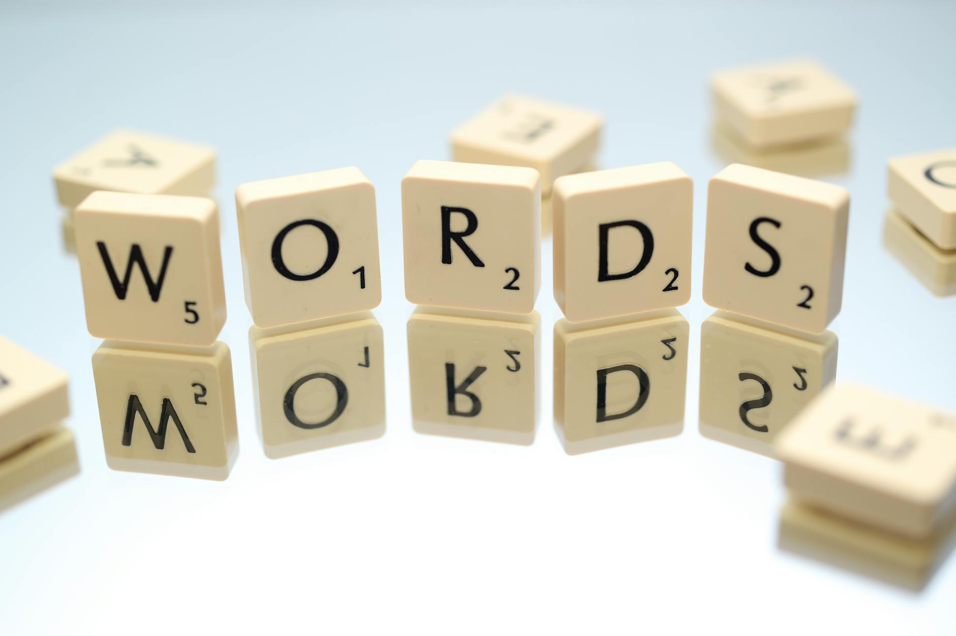 a series of scattered letter tiles, similar to those used in word-building board games like Scrabble. Each tile has a letter and a number, which typically represents the points the letter is worth in the game. The letters in the image are arranged to form the word "WORDS", with additional letters scattered around. The photo has a narrow depth of field, focusing on the "WORDS" in the centre while the other letters are blurred out in the foreground and background. The background is a light, solid colour, providing a clean, minimalistic look that highlights the tiles.