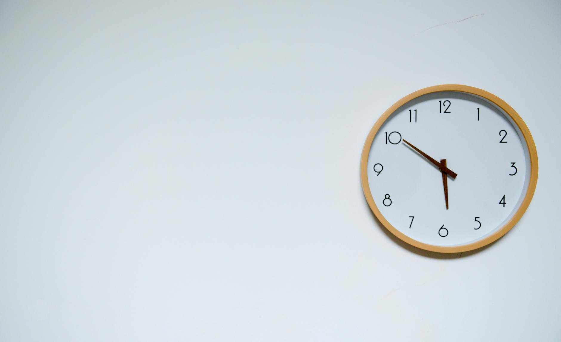 A round clock with black numbers on a white background hangs on a white wall. The time on the clock is ten to six.