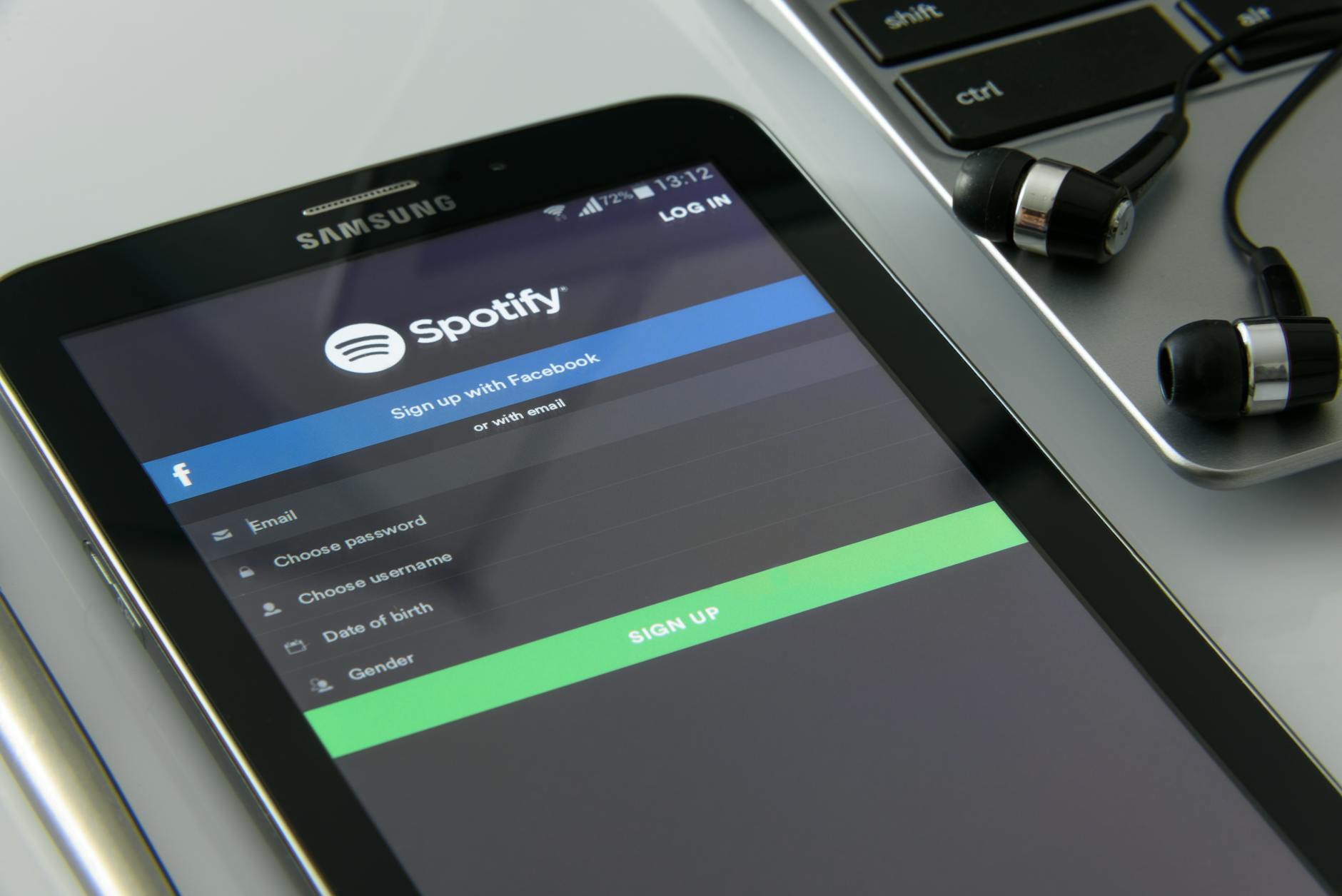 A Samsung tablet displaying the Spotify sign-up screen, next to a laptop and a pair of black earphones. The tablet screen shows options to sign up via Facebook or email, and input fields for account details.