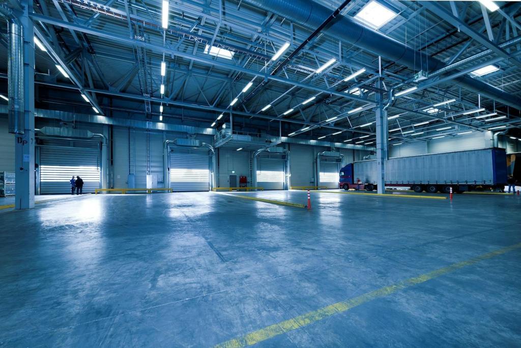 A spacious, well-lit warehouse interior with high ceilings, multiple loading docks, and a truck parked near one of the docks. Two people are standing in the distance near the loading bays.
