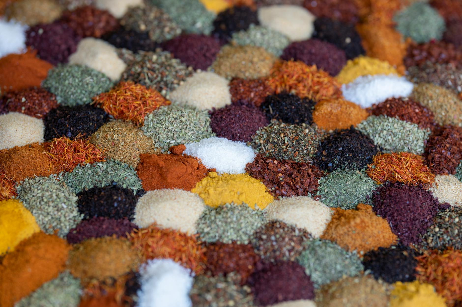 A close-up image of various colourful spices arranged in a grid pattern. The spices include different textures and colours, such as reds, greens, yellows, blacks, and whites, creating a vibrant mosaic.