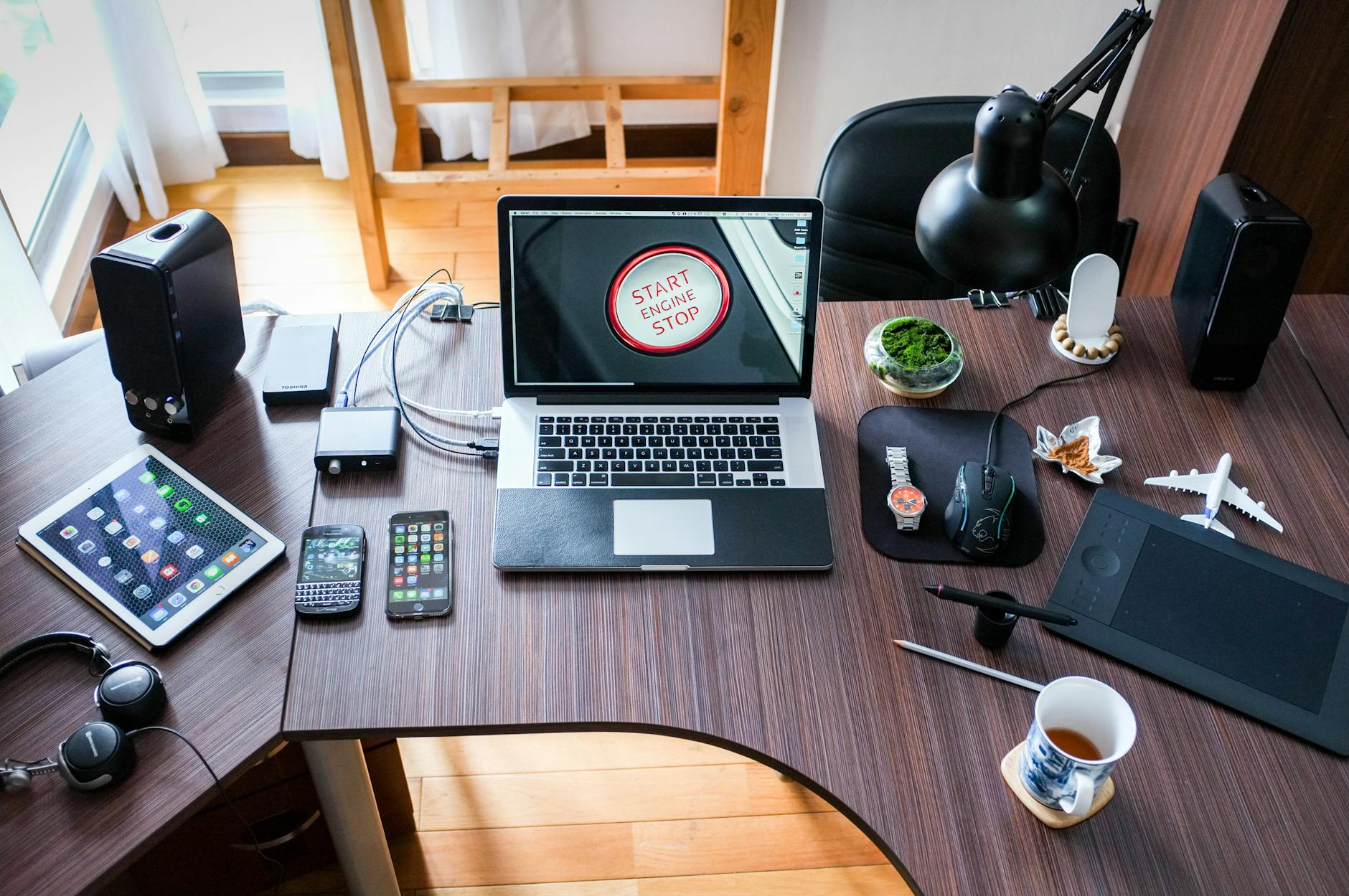 An image of a tech-filled workspace. There's a laptop with a "Start Engine Stop" button displayed on the screen, alongside multiple devices like tablets, phones, a drawing tablet, and various other tech gadgets. There's also a cup of tea, and a decorative airplane model on the table