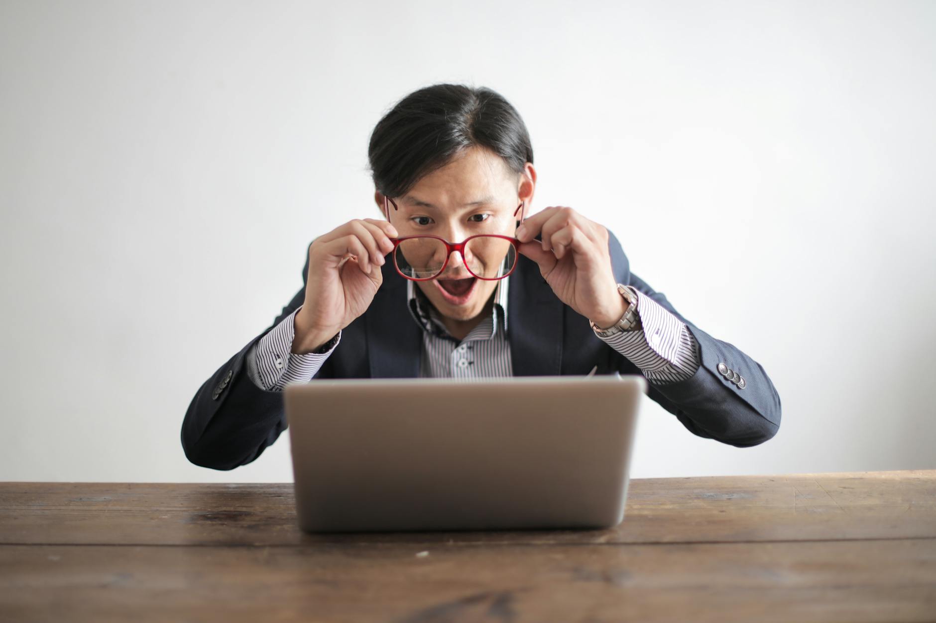 A surprised man wearing a suit and glasses is sitting at a wooden table, looking at a laptop screen. He is holding his glasses with both hands and has an expression of amazement or shock on his face.