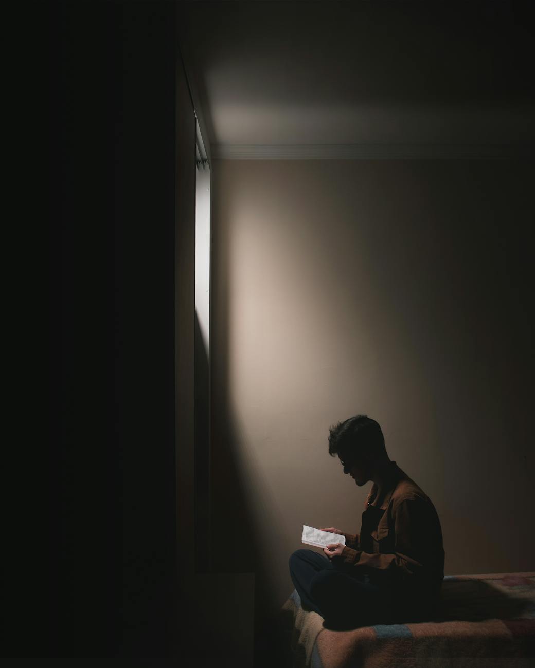 A person sits on a bed in a dimly lit room, illuminated by light from a window. The individual is reading a book, with their silhouette sharply contrasted against the light on the wall.
