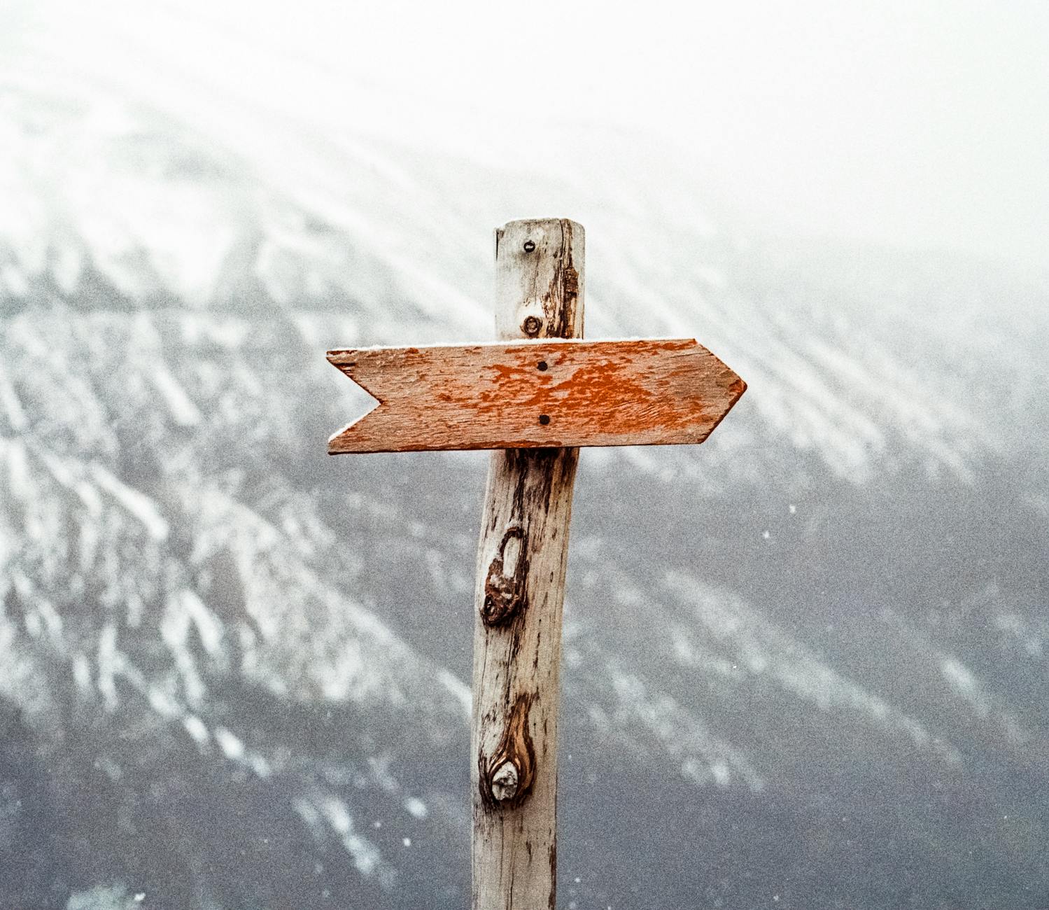 A wooden signpost with a single arrow pointing to the right, set against a snowy mountainous background. The sign has a rustic, weathered look, suggesting it has been exposed to the elements for some time. The background is somewhat blurred and covered in snow, contributing to a chilly, wintry atmosphere.