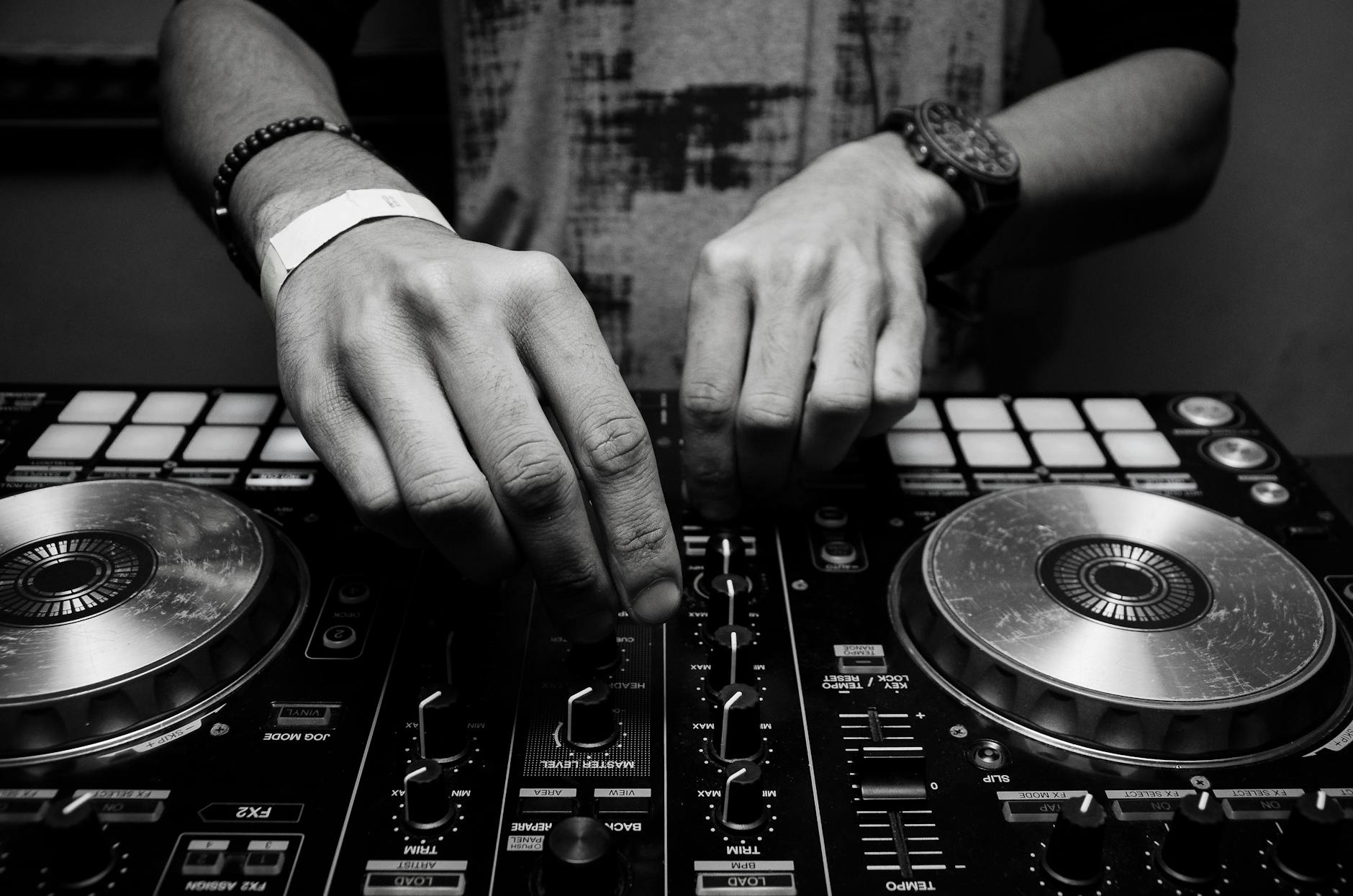 A black and white close-up of a DJ's hands operating a mixer, with fingers adjusting knobs and faders on the console. The DJ is wearing a wristband and a bracelet on one wrist, and a wristwatch on the other.