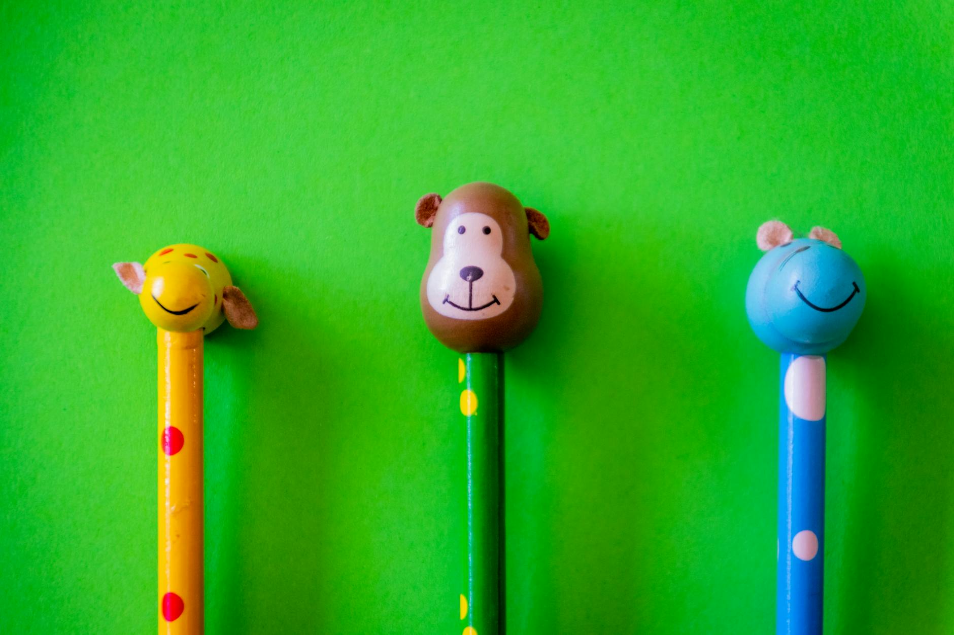 Three colorful pencils with animal head toppers are displayed against a bright green background. The left pencil has a yellow giraffe head, the middle pencil has a brown bear head, and the right pencil has a blue bear head.