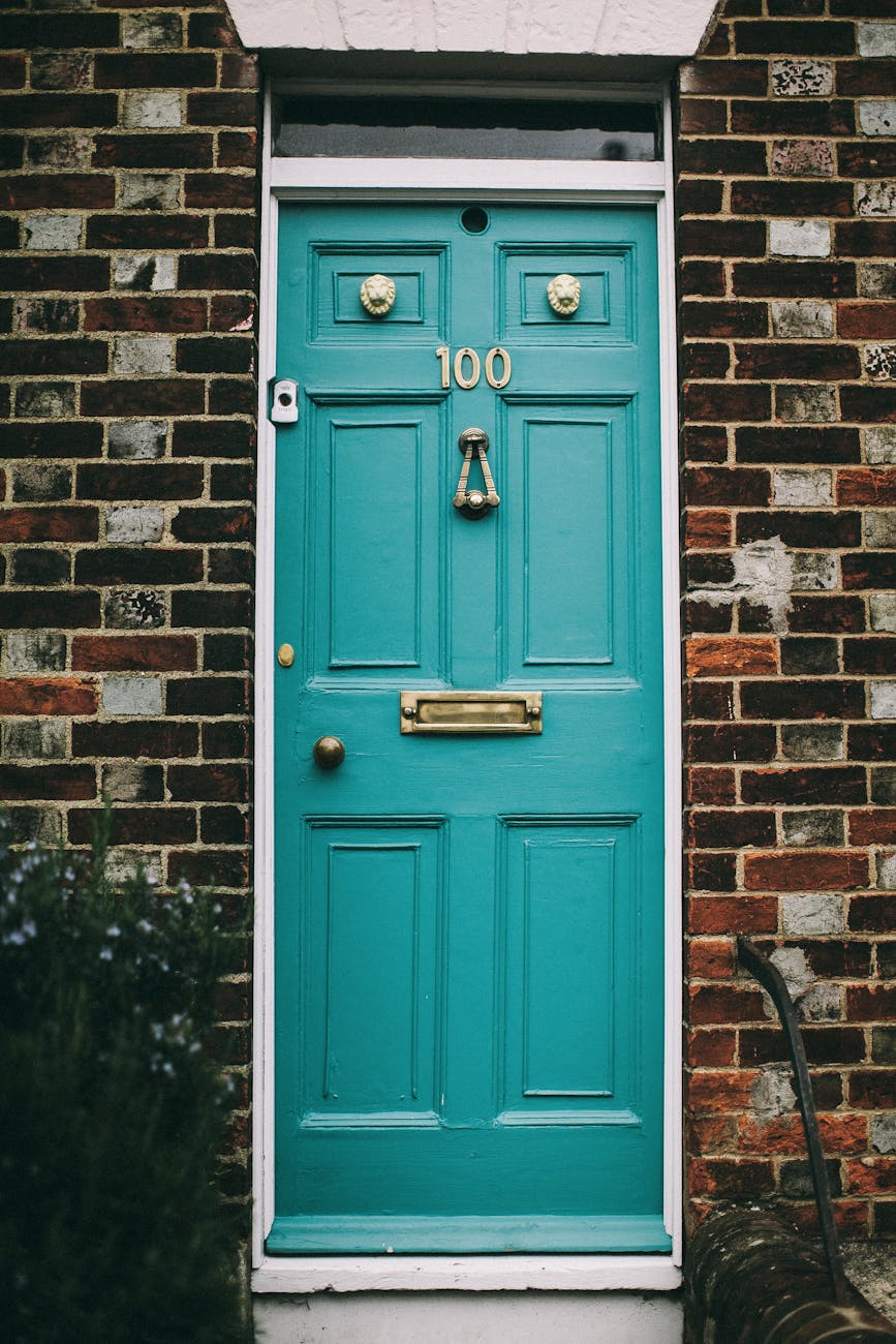 This is an image of a teal-colored door with the number "100" displayed prominently. The door has several brass fixtures including a knocker, a mail slot, and two lion head decorations. The door is set in a brick wall.