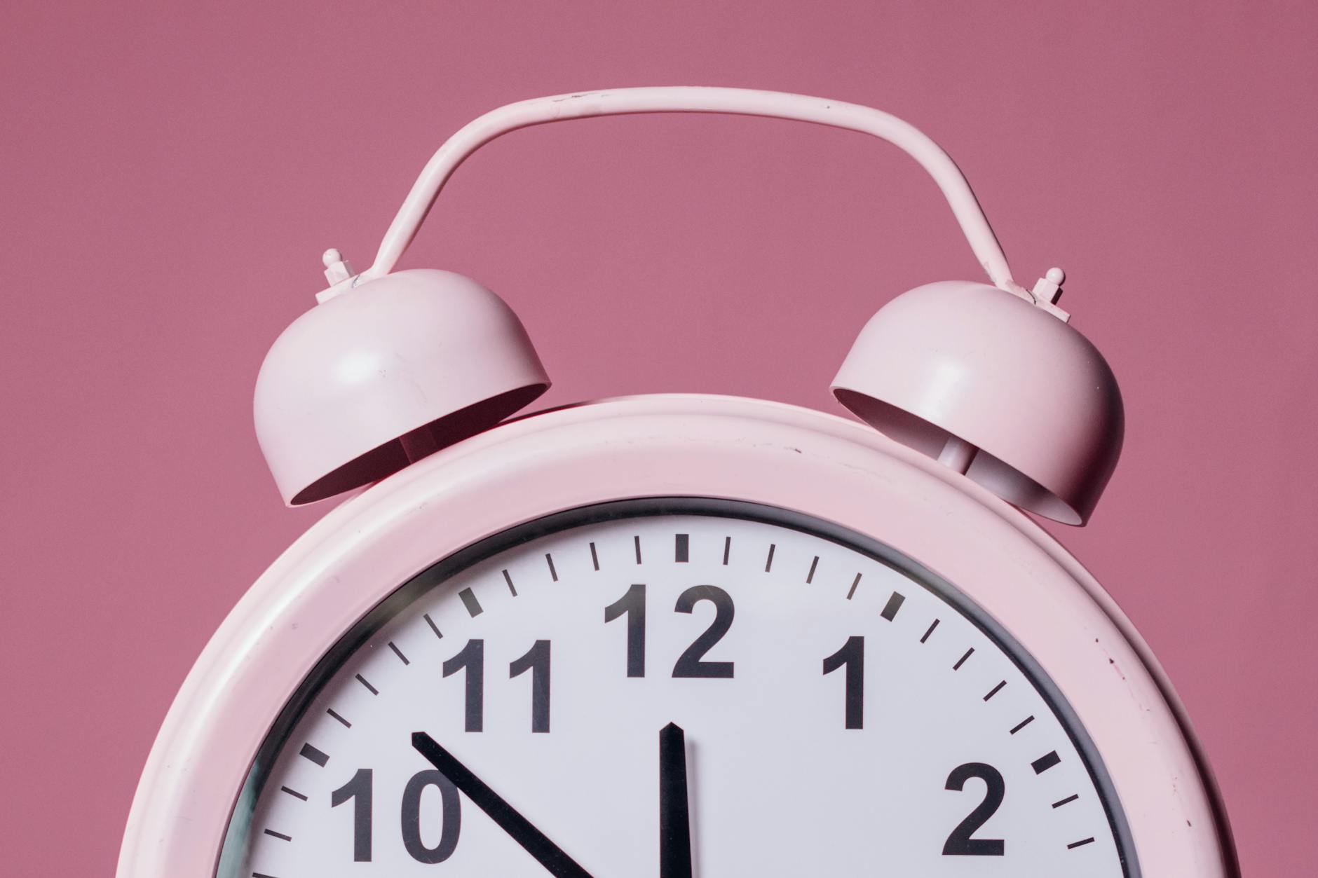 The image shows a close-up of a pink alarm clock with a white face and black numbers. The background is also pink. The clock hands are positioned near 10:10.