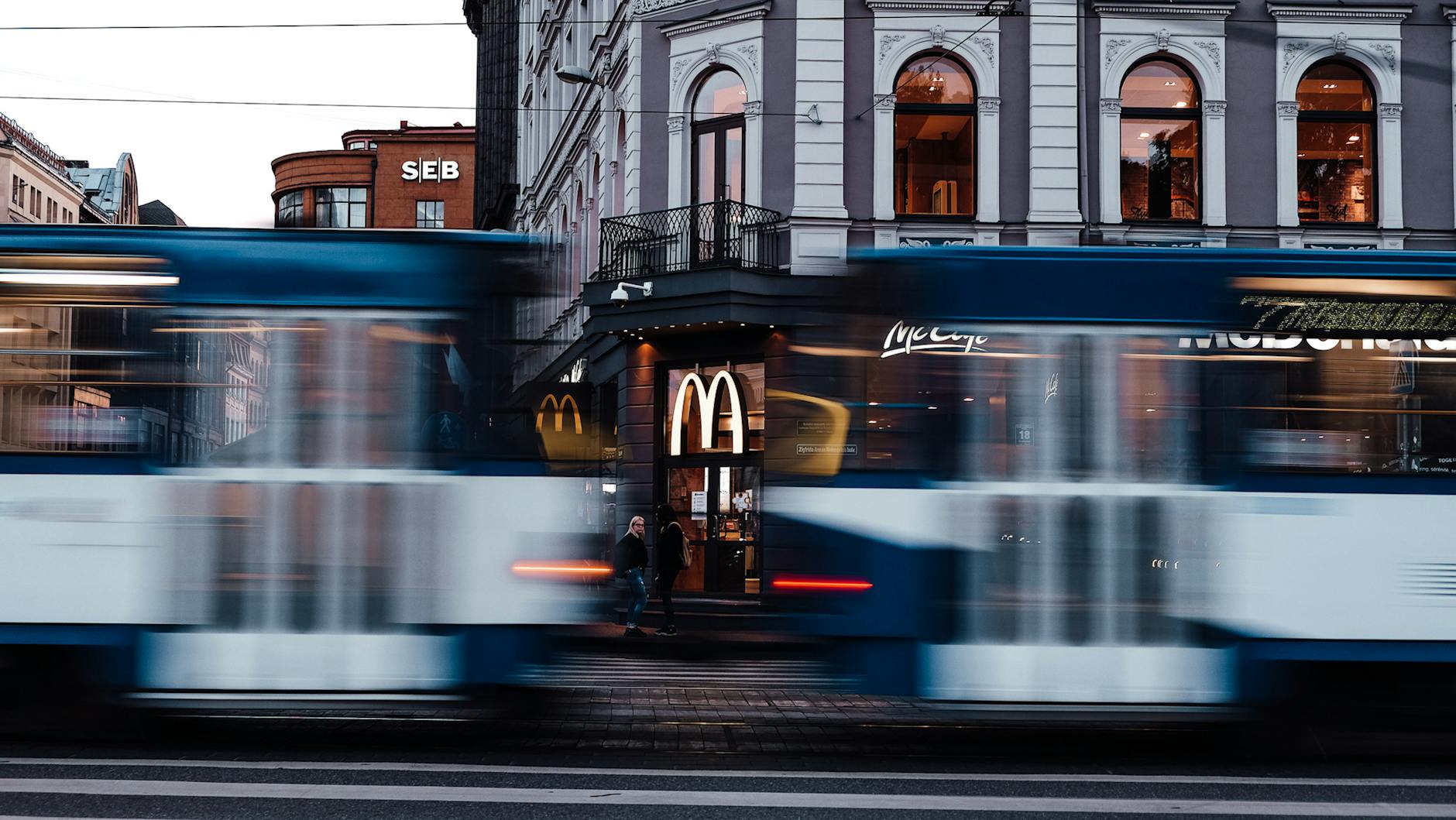 The image shows a bustling city street with a McDonald's restaurant, blurred tram cars, and a historic building with arched windows in the background.

