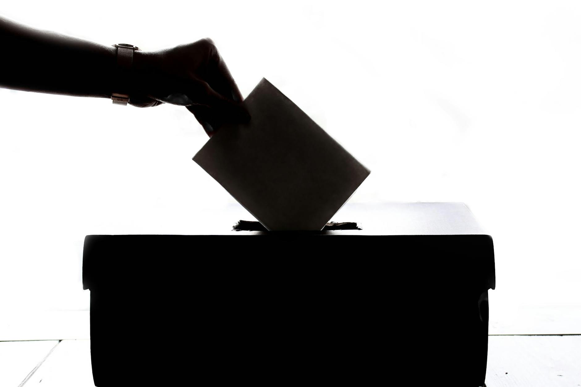 The image shows a hand placing a ballot into a ballot box, representing the act of voting in an election.