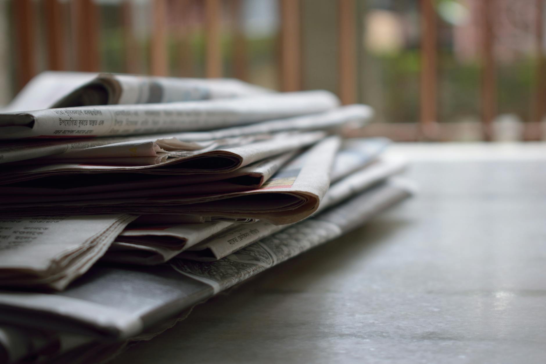 Stacked newspapers waiting to be read on a table, with a blurred outdoor environment visible in the background.

