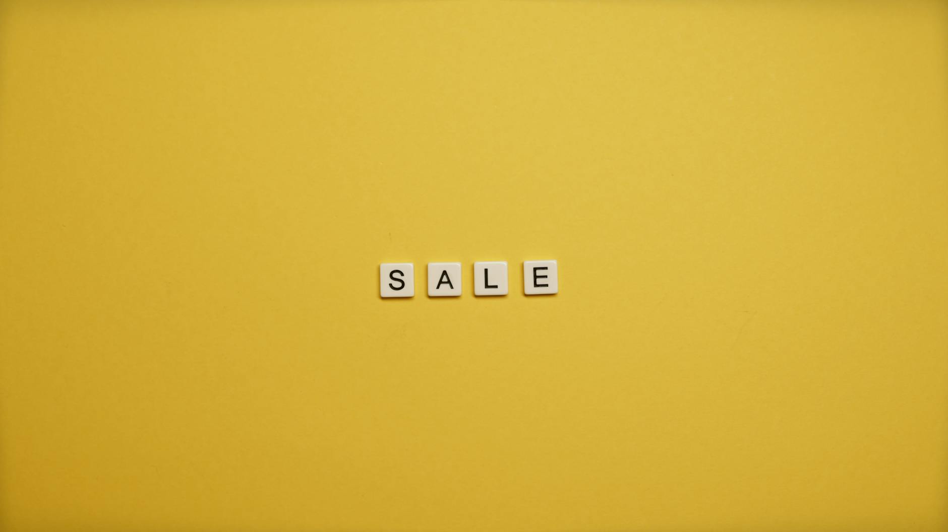 The image shows the word "SALE" spelled out using scrabble-style letter tiles on a bright yellow background.