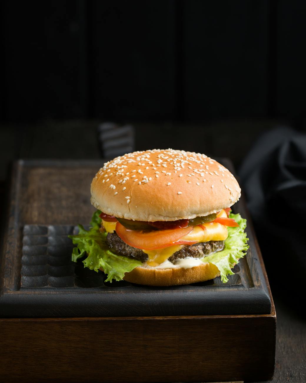A close-up image of a cheeseburger with a sesame seed bun, fresh lettuce, tomato slices, melted cheese, pickles, and a beef patty, placed on a dark wooden tray against a dark background.