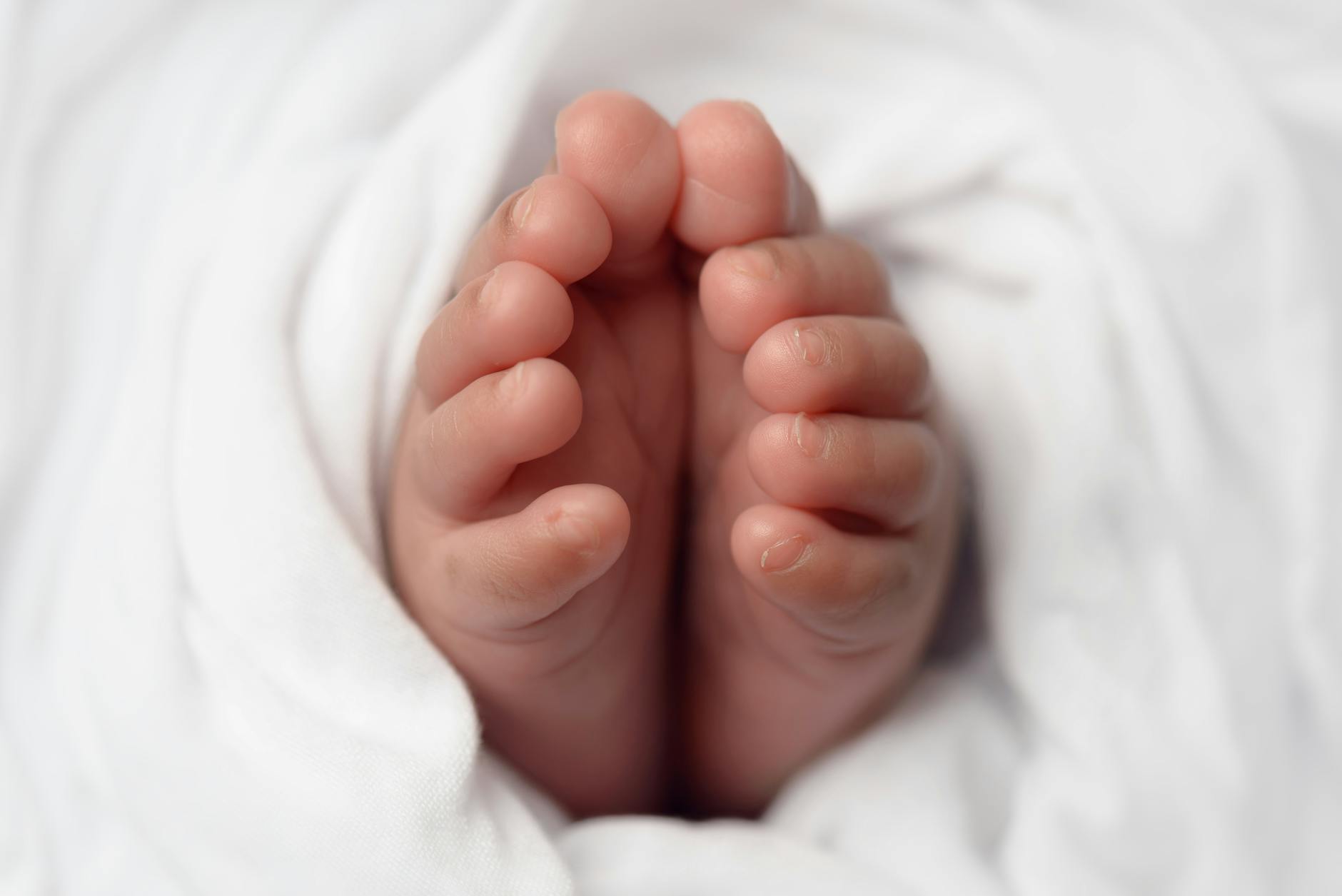 A close up photo of a baby's feet, with all ten toes showing. The baby is wrapped in a white cotton sheet.