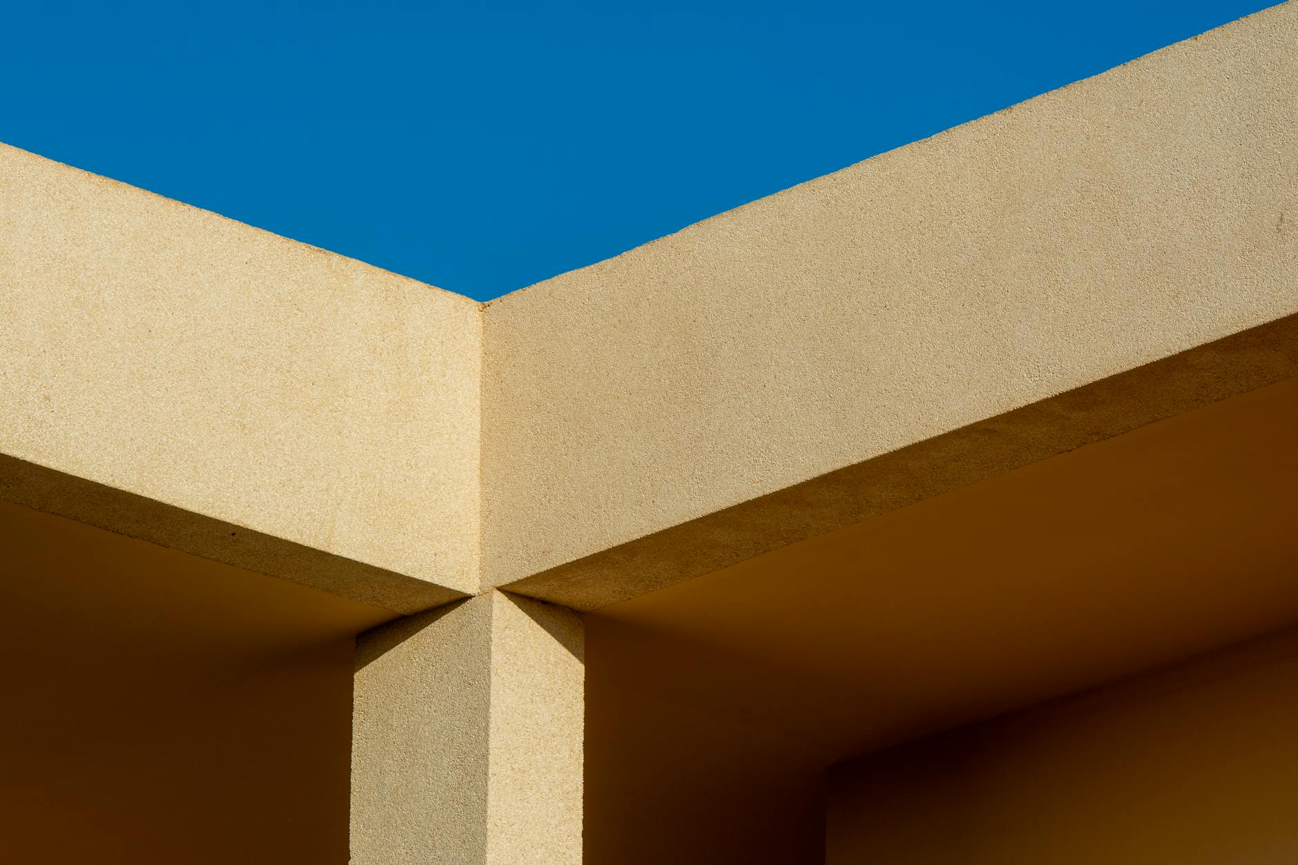 Minimalist architecture featuring intersecting beige walls and shadows against a clear blue sky.