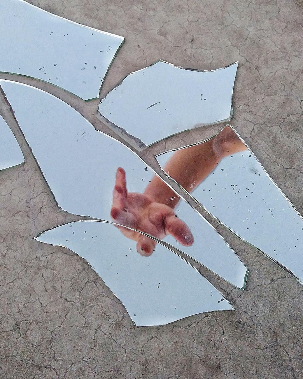 A close-up view of broken mirror shards scattered on a surface, with a hand reaching through them. 