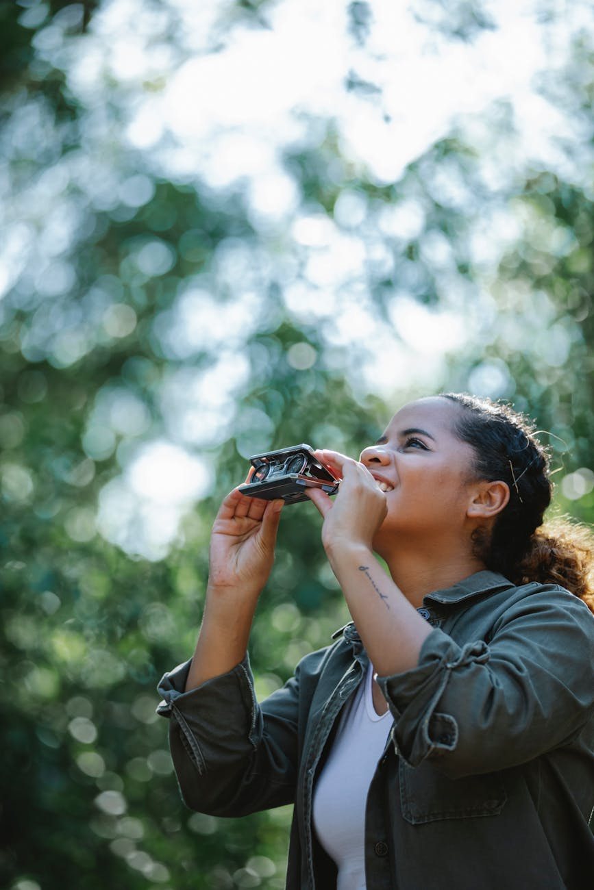 A woman looks up while holding a pai of folding binoculars, surrounded by blurred greenery and dappled sunlight, conveying a sense of exploration and joy.