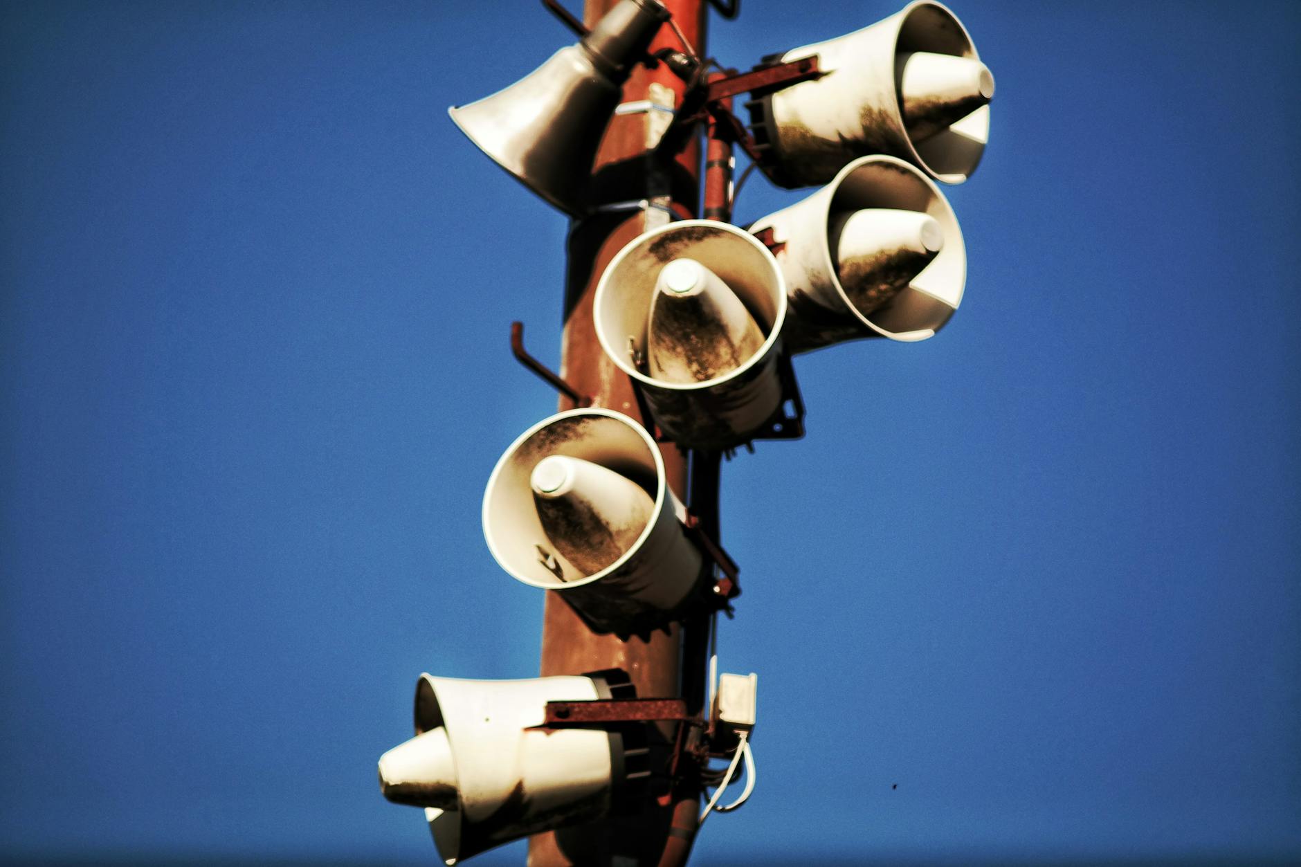 A cluster of weathered loudspeakers mounted on a pole against a clear blue sky.