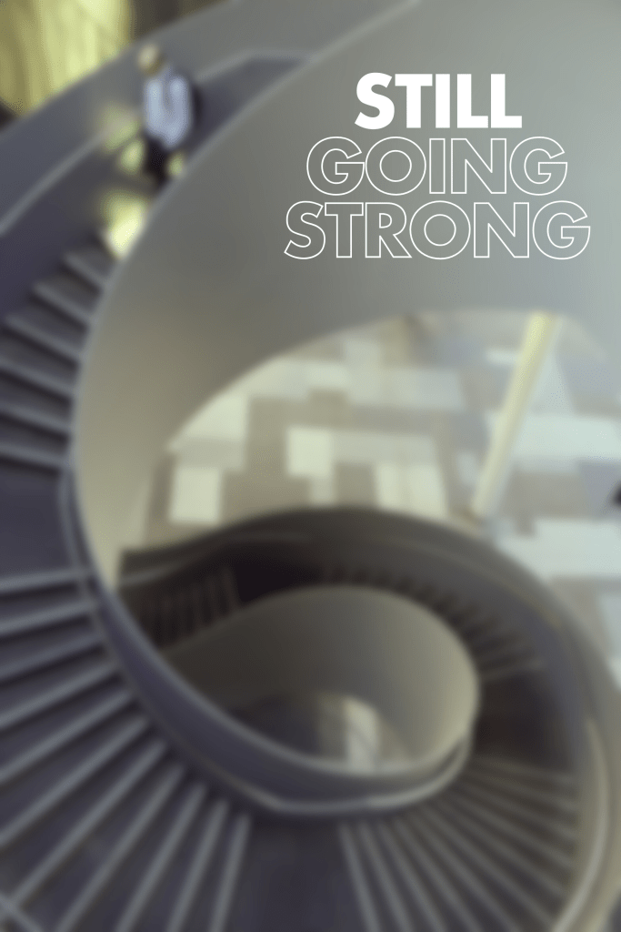 "Text 'STILL GOING STRONG' with a blurred image of a spiral staircase and a person walking"

