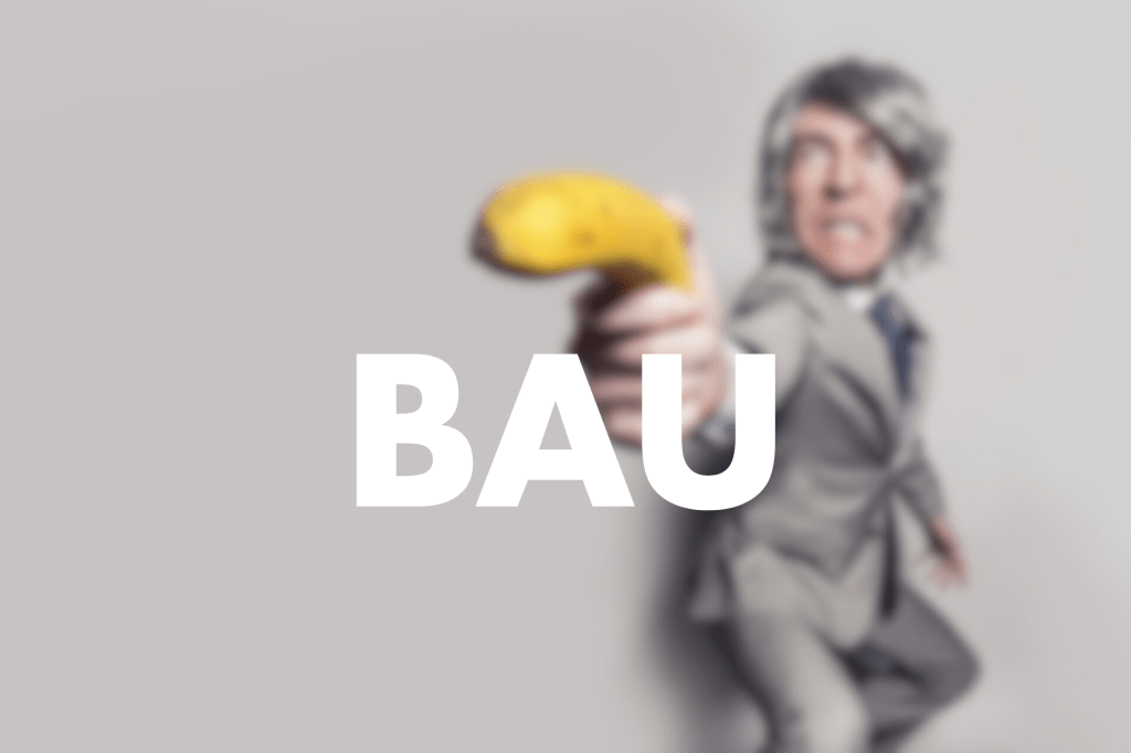 The image features a person in a suit dramatically pointing a banana, with the word "BAU" prominently displayed in bold white text across the foreground. The background is blurred, emphasising the action and the text.
