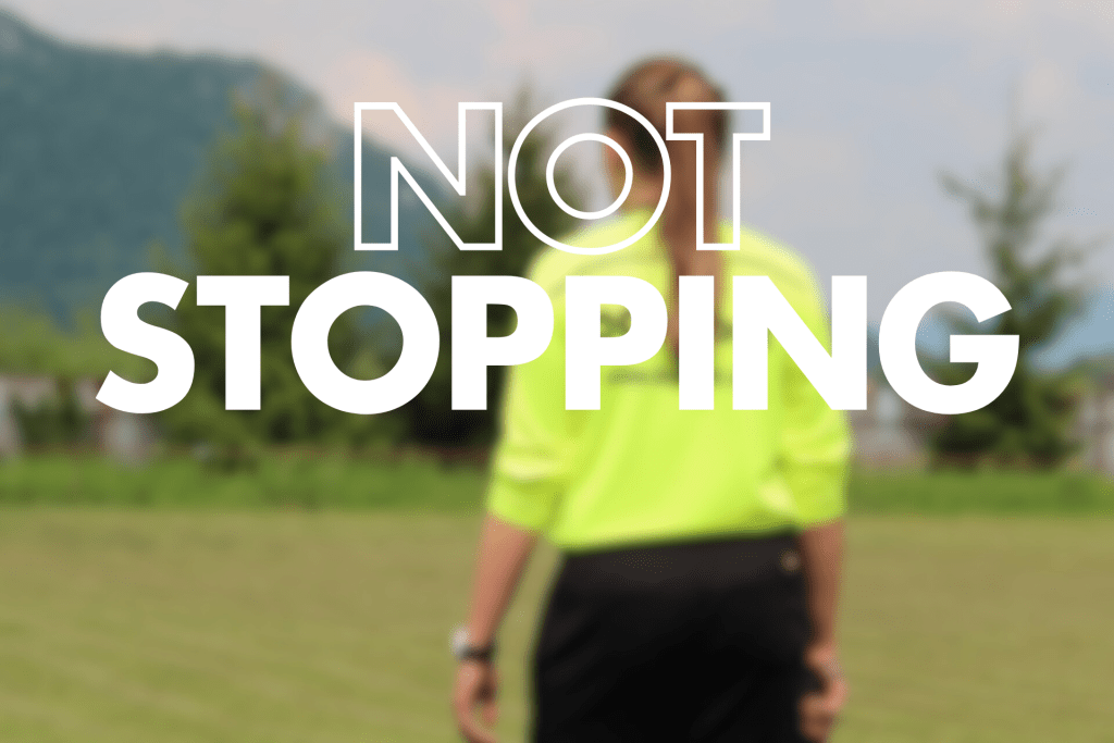 "NOT STOPPING" text overlays a blurred image of a female assistant referee in a bright yellow top and dark shorts, standing in a grassy field.