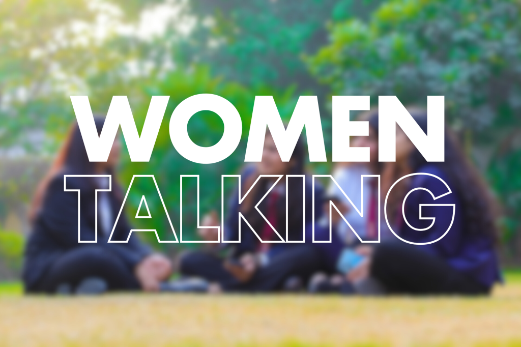 A group of women sits together on the grass, engaged in conversation. Bold text overlays the image, reading "WOMEN TALKING," highlighting their interaction.

