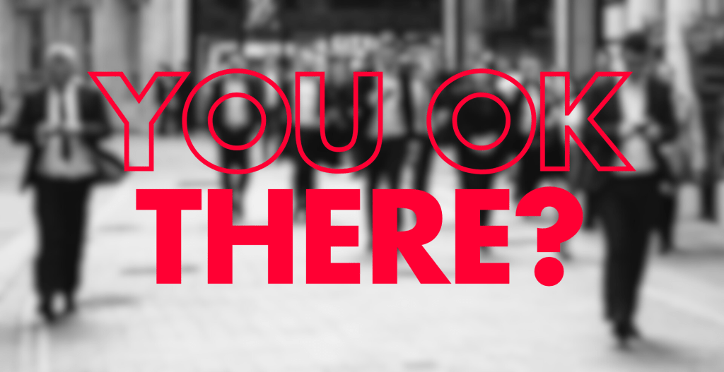 This image shows bold red text saying "YOU OK THERE?" overlaid on a blurry black and white background. The background appears to show people walking holding mobile phones in what might be a trade show, exhibition hall, or busy public space.