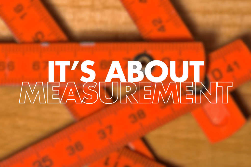 The image shows several orange measuring tapes or rulers against a wooden background. Overlaid on the image is text that reads "IT'S ABOUT MEASUREMENT" in large white letters. The word "MEASUREMENT" appears to be in an outline font.
