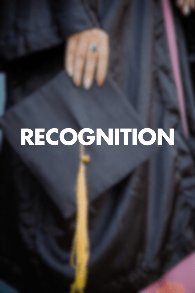 The image shows the word "RECOGNITION" in large white text overlaid on a blurred background featuring a graduation cap with a yellow tassel. A hand is visible at the top of the frame, appearing to hold or adjust the cap. The person is wearing what seems to be a dark graduation gown, suggesting an academic achievement or commencement context.