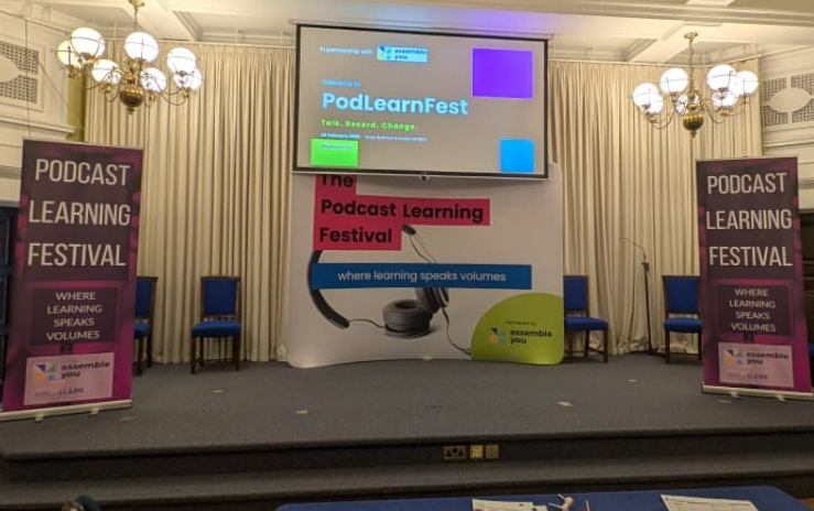 A lecture stage with a central screen reading "PodLearnFest," flanked by two vertical banners that say "Podcast Learning Festival." A backdrop shows oversized headphones and the tagline "where learning speaks volumes."